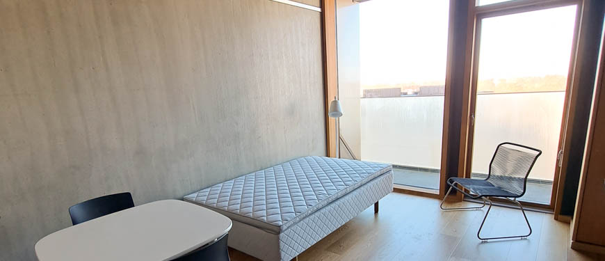 Billedet viser lejlighedens opholdsrum med spisekrog og sovesofa op ad højre væg, skrivebord og reoler på venstre væg, og panoramavindue med udgang til balkonen bagerst i billedet.
