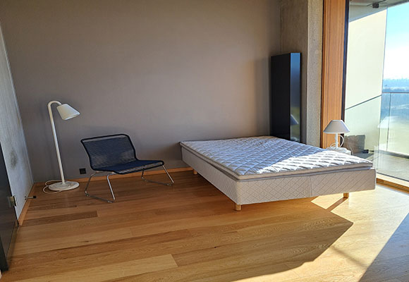 The image shows the living area. To the left is the door to the bathroom. In the centre is a sofa bed, lamp and chair and to the right is the panoramic view and exit to the balcony.