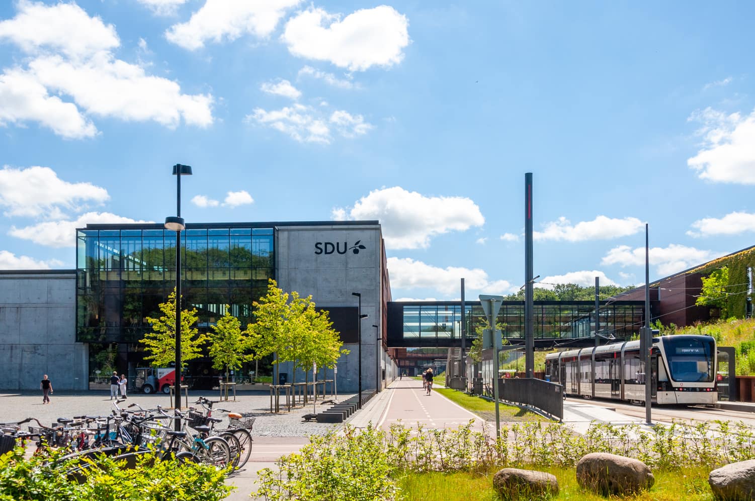 SDU-campus med glasbygning, cykelparkering og letbane på en solrig dag.