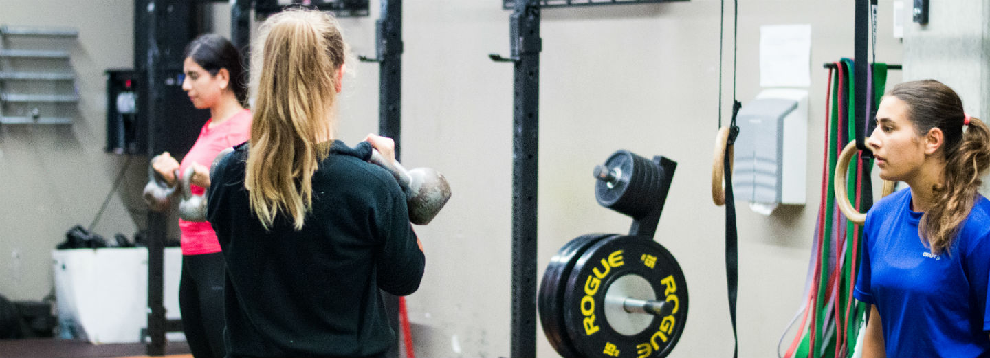 Tre kvinder laver træning i et fitnesscenter med kettlebells, "ROGUE"-vægtskiver, gymnastikringe og elastikker.