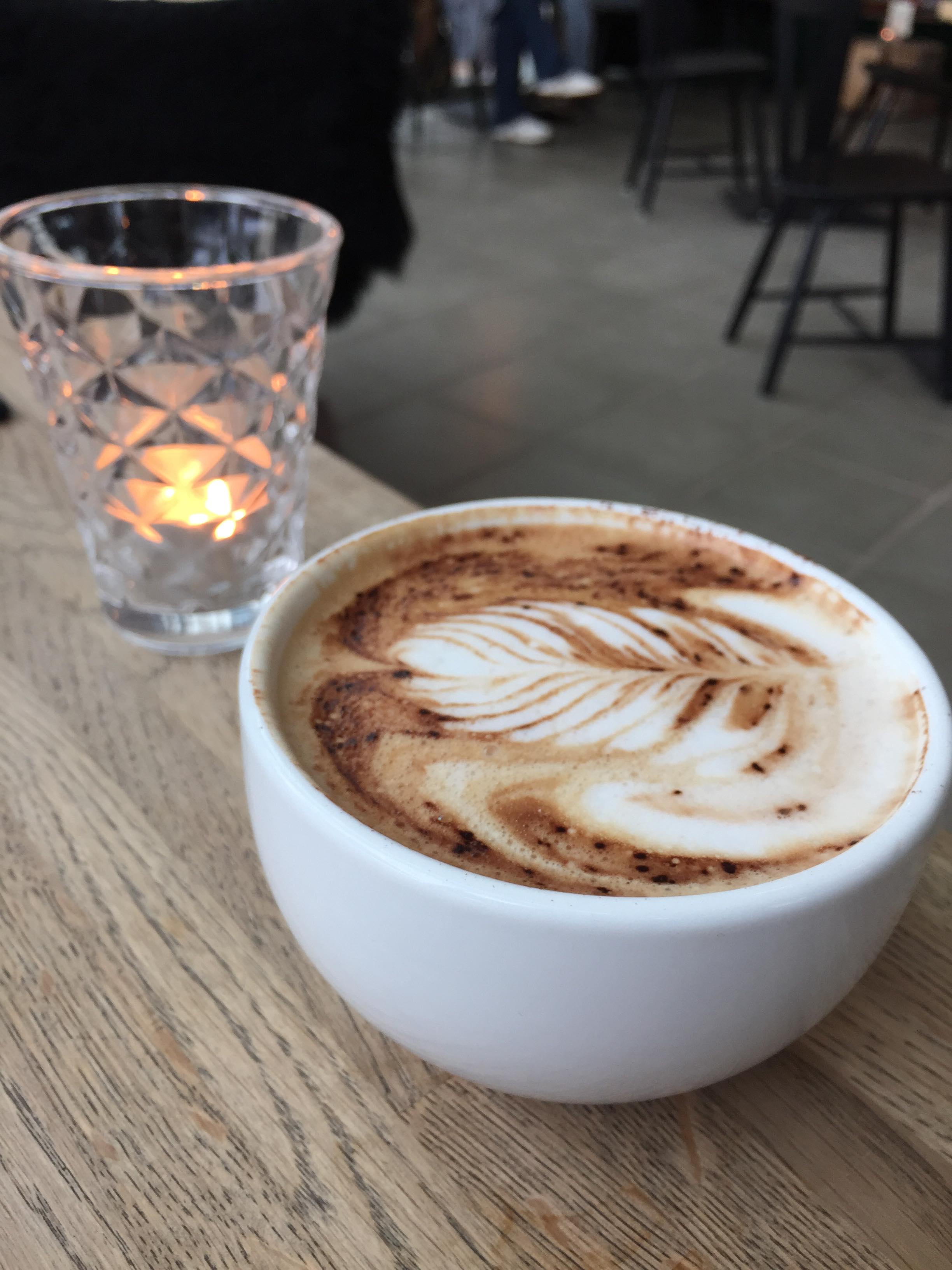 Hvid kaffekop med lattekunst på et træbord ved siden af et glas med tændt lys i en café.